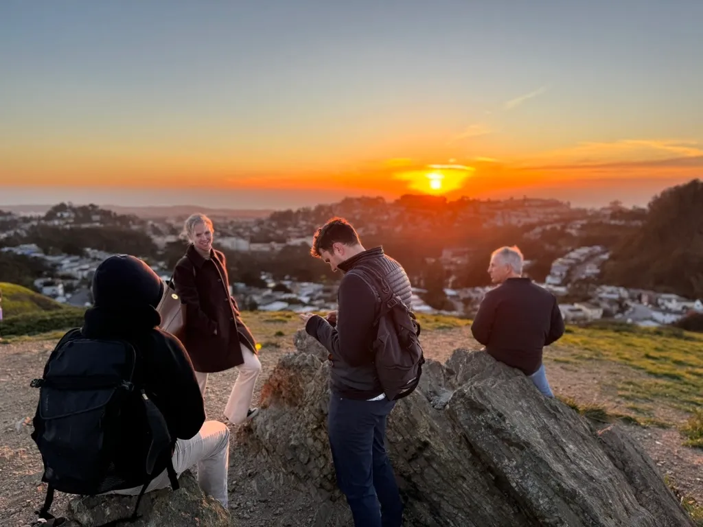 Four people sitting and standing on rocks overlooking a city at sunset